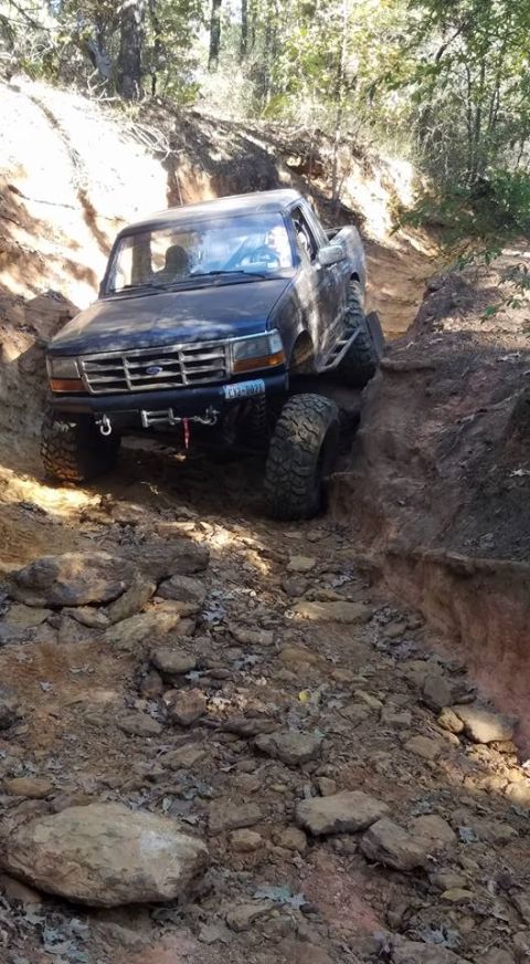 1995 Ford Bronco - A dark blue Ford pickup truck is navigating a steep, rocky, and muddy off-road trail. The front of the truck is angled towards the viewer,…