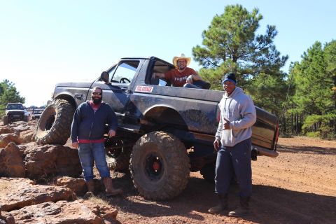 1995 Ford Bronco - Three men pose with a heavily modified black Ford pickup truck with oversized tires, parked on a dirt trail with rocks and trees in the bac…