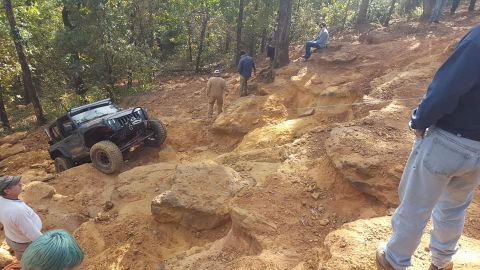 1995 Ford Bronco - A black Jeep Wrangler is navigating a rocky, off-road trail with several people watching and assisting. The vehicle is angled upwards on a…
