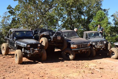 1995 Ford Bronco - Several off-road modified vehicles are parked on a dirt incline with trees in the background. A black Ford Bronco is in the center, flanked…