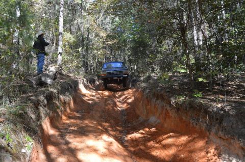 1995 Ford Bronco - A dark blue Ford Bronco navigates a deep, rutted dirt trail through a wooded area. A person wearing a cowboy hat stands on the embankment t…