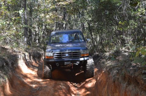1995 Ford Bronco - A dark blue 1995 Ford Bronco is driving through a narrow, rutted dirt trail in a wooded area. The front of the vehicle is visible, showing…