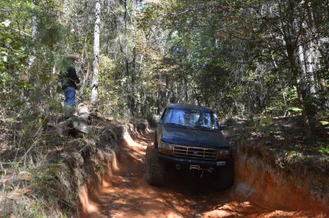1995 Ford Bronco - A dark-colored Ford Bronco navigates a deep, rutted dirt trail through a dense forest. A person wearing a hat stands on the embankment to t…