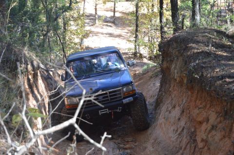 1995 Ford Bronco - A dark blue Ford Bronco navigates a narrow, rutted dirt trail with steep, eroded banks on either side. The vehicle is angled sharply, with…