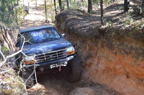 1995 Ford Bronco - A dark blue Ford Bronco navigates a narrow, rutted dirt path with steep, eroded earthen walls on either side. The vehicle is angled towards…