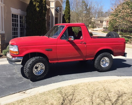 1994 Ford Bronco - A red 1990s Ford Bronco is parked on a paved driveway in front of a house. The two-door SUV has large off-road tires and a spare tire mount…