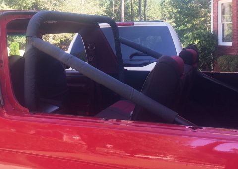 1994 Ford Bronco - The interior of a red 1994 Ford Bronco is shown, featuring bucket seats and a roll cage with padded bars. A white SUV is visible behind the…