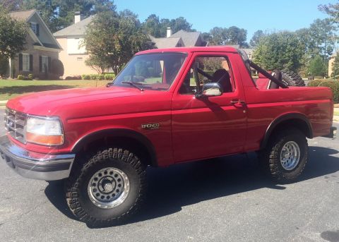 1994 Ford Bronco - A red 1994 Ford Bronco is parked on an asphalt surface in front of residential homes. The vehicle is a two-door model with a roll bar and a…