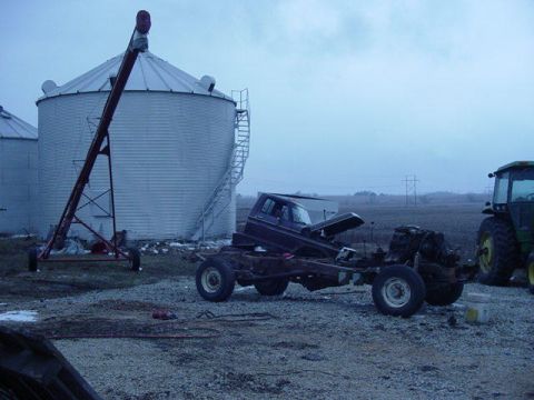 1992 Ford Bronco - Truck chassis and cab sit outside near grain silos and a tractor on a cloudy day.