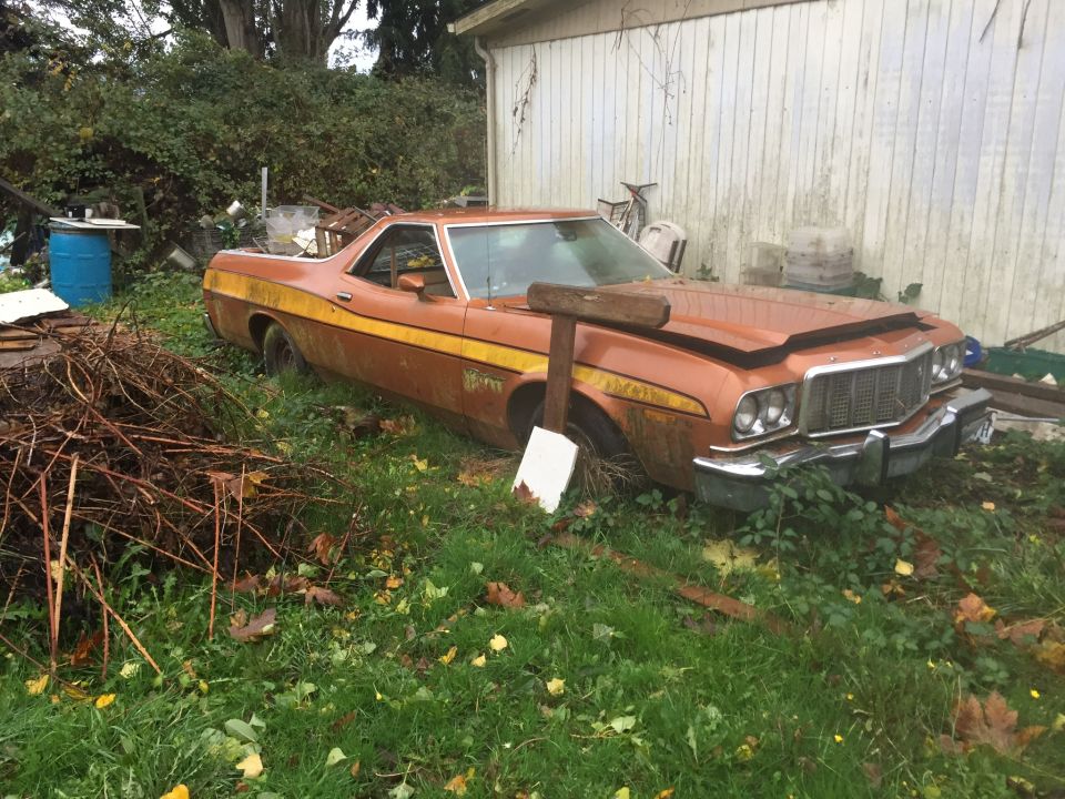 Rusty orange Ford Ranchero with yellow stripes parked in overgrown grass next to a white barn