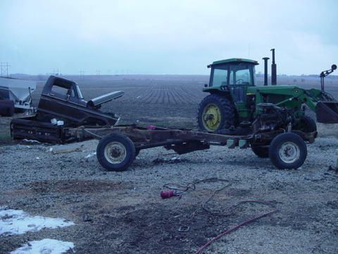 1992 Ford Bronco - A full-size truck/SUV body sits stripped off its frame in a gravel farm lot, the bare rusty ladder chassis with axles and wheels rolled out…