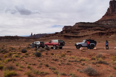 1995 Ford Bronco - Three off-road vehicles are parked on a dirt road in a desert landscape with red rock formations in the background. A red Ford Bronco is pa…