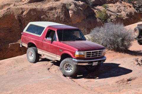 1995 Ford Bronco - A red 1995 Ford Bronco with a white roof is parked on a rocky, desert terrain. The SUV is angled towards the viewer's left, showcasing its…