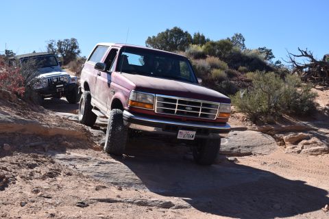 1995 Ford Bronco - A maroon 1995 Ford Bronco is driving on a rocky, off-road trail. Another silver Jeep is visible behind it.