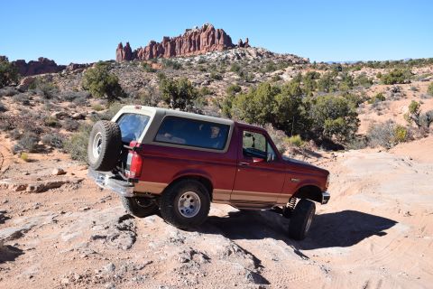 1995 Ford Bronco - A red 1995 Ford Bronco is shown off-roading on a rocky, desert terrain with red rock formations in the background.
