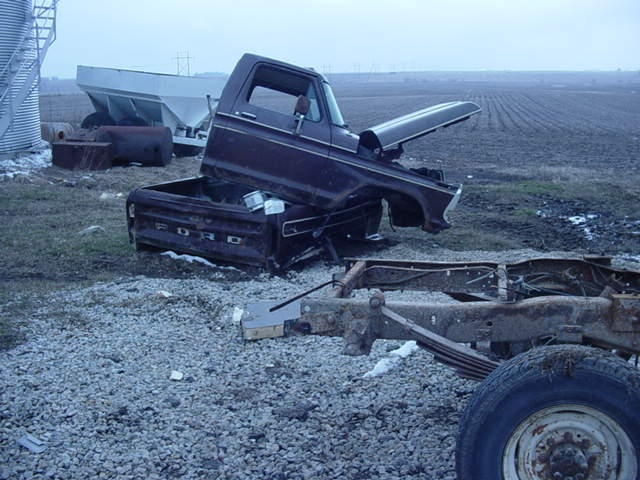 Dark red Ford truck cab and bed sitting on gravel, hood open. Chassis and front wheel visible in foreground