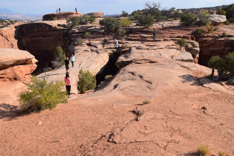1995 Ford Bronco - A group of people are walking on a rocky, arid landscape with red dirt and sparse vegetation. A deep crevice is visible in the center of th…