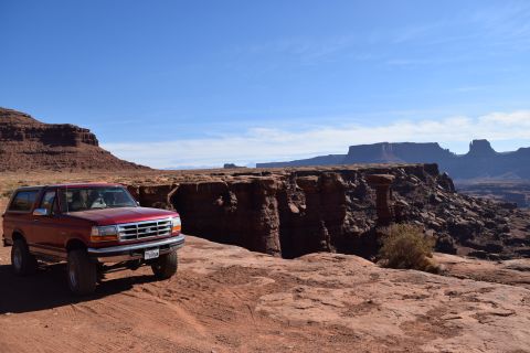 1995 Ford Bronco - A red 1995 Ford Bronco is parked on a dirt road overlooking a vast canyon landscape with red rock formations and a clear blue sky.