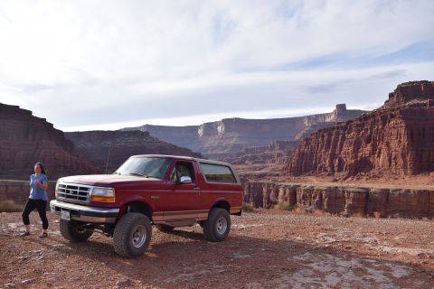 1995 Ford Bronco - A red 1995 Ford Bronco is parked on a dirt road with a woman standing next to it. The vehicle is positioned in front of a vast canyon lands…