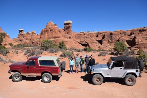 1995 Ford Bronco - A red 1995 Ford Bronco with a tan top and a silver Jeep Wrangler are parked on a dirt road in a desert landscape with red rock formations u…