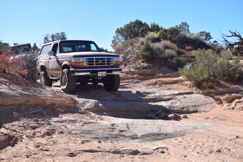 1995 Ford Bronco - A maroon 1995 Ford Bronco is shown off-roading on a rocky, desert trail under a clear blue sky. The vehicle is positioned at an angle, navi…