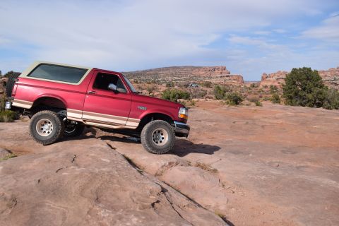 1995 Ford Bronco - A red 1995 Ford Bronco is parked on a rocky, desert-like terrain with mesas and sparse vegetation in the background under a partly cloudy s…
