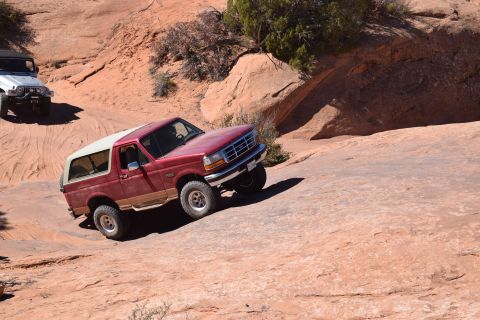 1995 Ford Bronco - A red 1995 Ford Bronco with a white removable top is driving off-road on a rocky, sandy terrain. Another white Jeep is visible in the backg…
