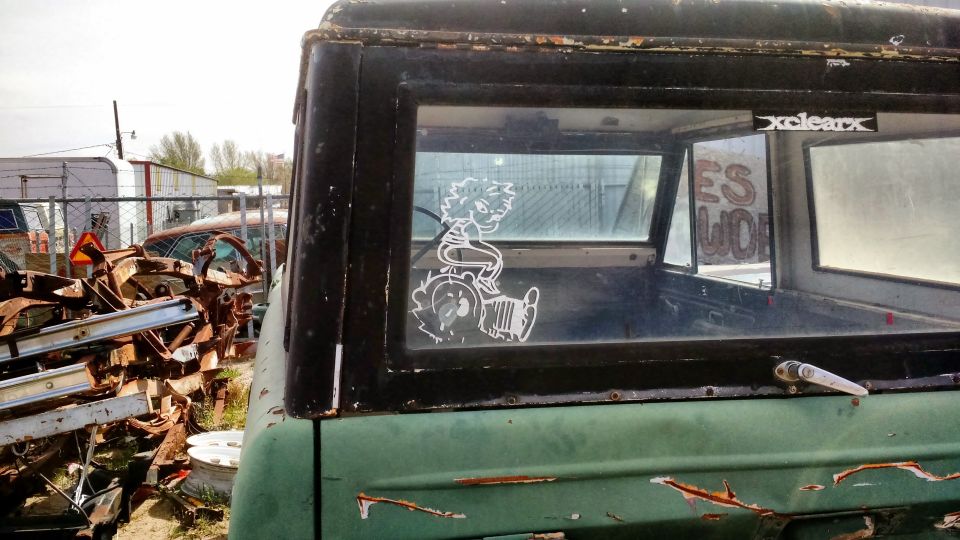Weathered green 1971 Ford Bronco in a junkyard with a cartoon sticker on the rear window.
