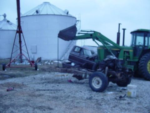 1992 Ford Bronco - A tractor loader is lifting the chassis of a brown Ford Bronco, which appears to be from the 1980s, in front of large silos.