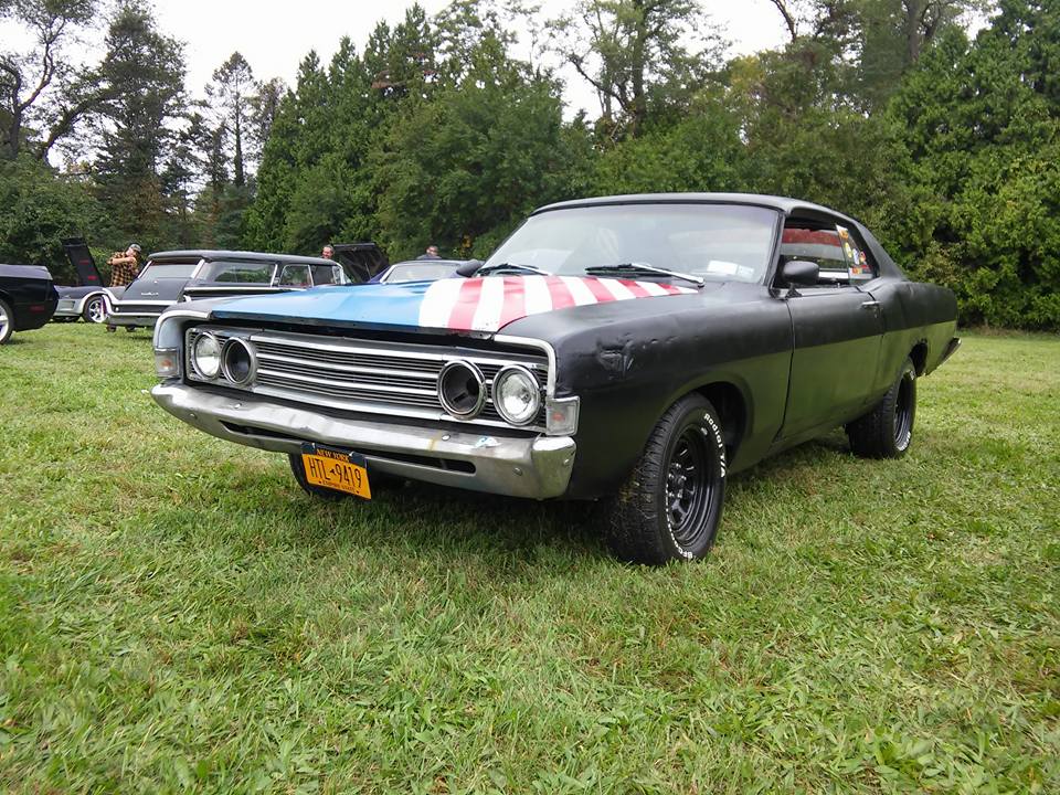 Black 1969 Ford Torino with American flag on hood, parked on grass.