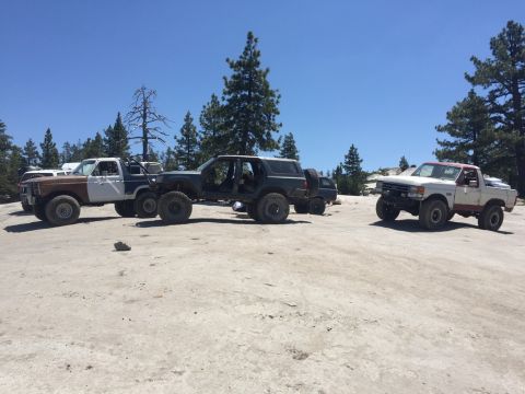 1984 Ford Bronco - Three modified off-road vehicles are parked on a rocky, dirt surface under a clear blue sky. The vehicles include a two-tone Ford Bronco, a…