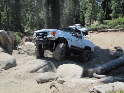 1984 Ford Bronco - A white and maroon Ford Bronco is navigating a rocky, off-road trail in a forest. The vehicle appears to have some damage to its front end…