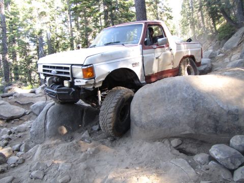 1984 Ford Bronco - A white and red Ford pickup truck is navigating a rocky, off-road trail, with its front tire perched precariously on a large boulder. The t…