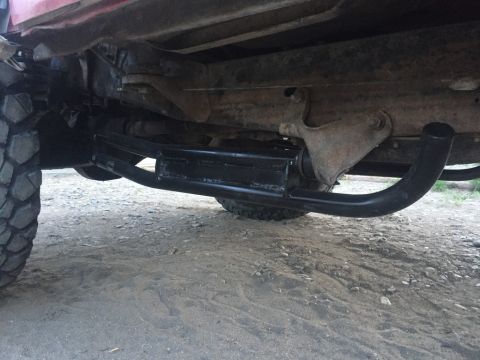 1984 Ford Bronco - The underside of a red Ford Bronco shows a black steel step bar installed below the frame, extending from the front wheel well area towards…