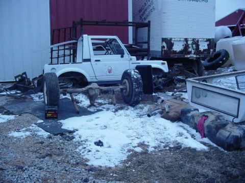 1992 Ford Bronco - A white Suzuki Samurai is partially disassembled and sits on a chassis in a snowy yard with various scrap metal parts around it.
