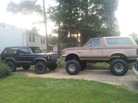 1988 Ford Bronco - Two lifted Ford Broncos are parked side-by-side in a driveway with a house and trees in the background. The Bronco on the left is black, an…
