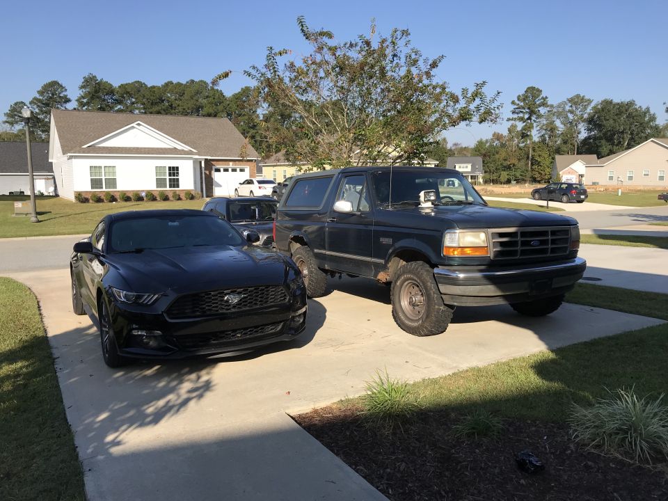 1990s dark blue Ford Bronco parked next to a black Ford Mustang in a driveway.