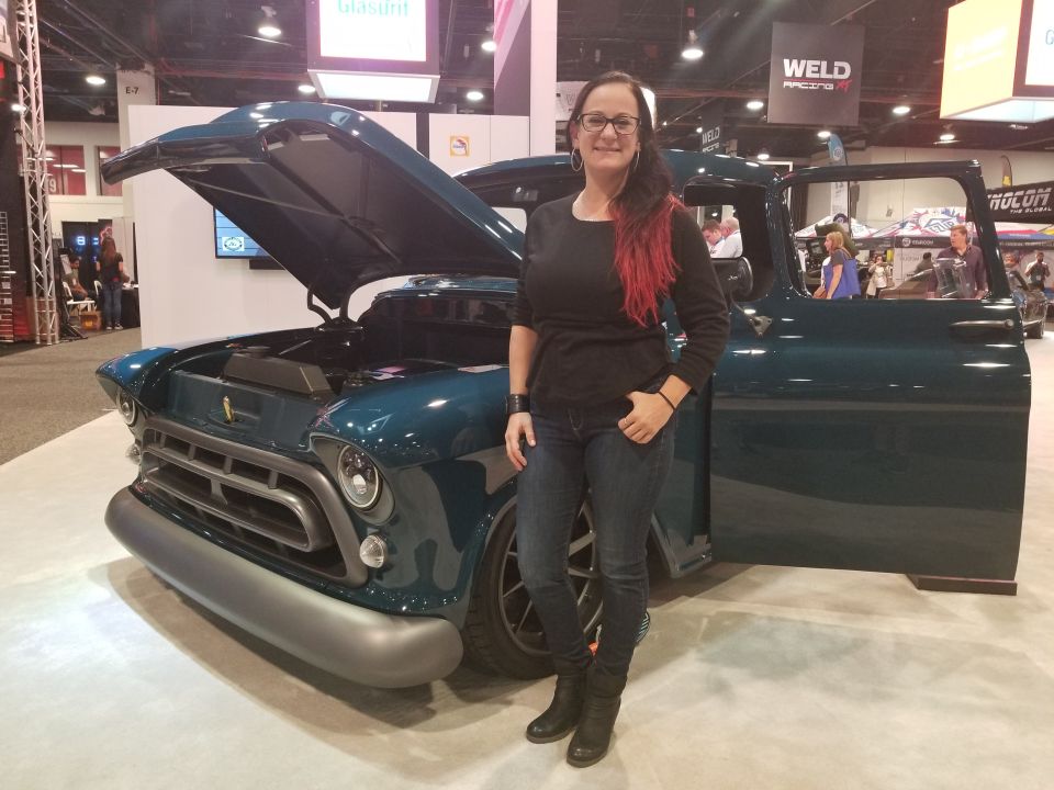 Woman standing next to a vintage dark teal pickup truck with hood and door open.