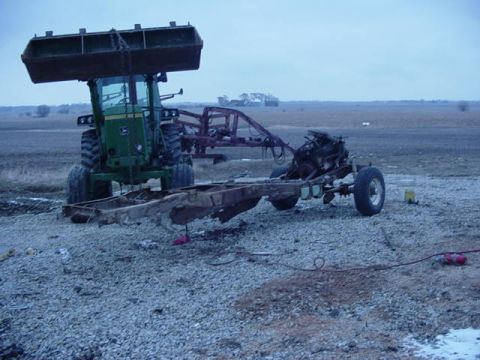1992 Ford Bronco - A tractor lifts a vehicle frame with its engine and drivetrain, suggesting a dismantling or salvage operation.
