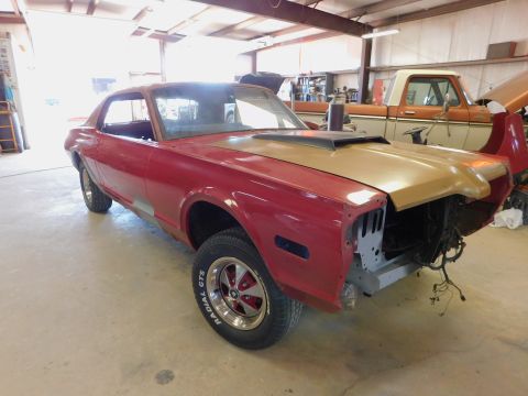 1968 Mercury Cougar - A red coupe with a gold hood and scoop is in a garage, missing its front end, with an orange pickup truck behind it.