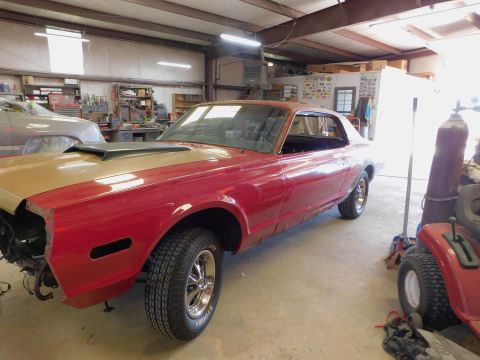 1968 Mercury Cougar - A red and gold 1968 Mercury Cougar coupe is in a garage, appearing to be under repair, with another vehicle and equipment visible.