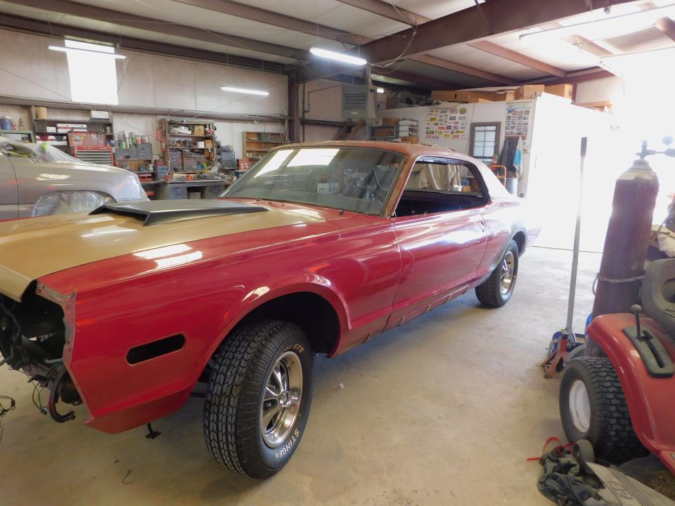 Red and gold 1968 Mercury Cougar coupe with a hood scoop, missing front bumper, in a garage