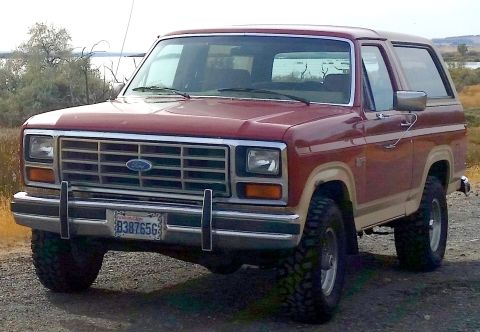 1985 Ford Bronco - A red and tan 1980s Ford Bronco is parked outdoors on a dirt road with a body of water and dry brush in the background. The vehicle has a c…
