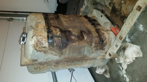 1985 Ford Bronco - A heavily rusted and corroded fuel tank is shown removed from a vehicle, resting on the ground next to a garage door. The tank appears to b…