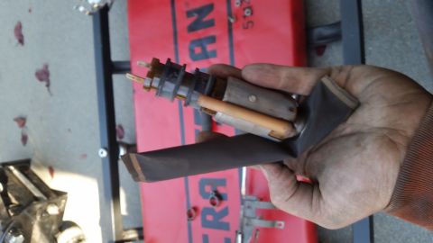 1985 Ford Bronco - A hand holds a disassembled fuel pump assembly, likely from a 1985 Ford Bronco, against a red mechanic's creeper. The part includes a black…