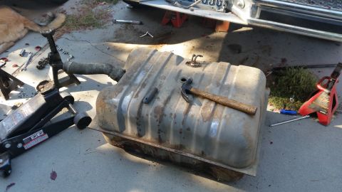 1985 Ford Bronco - A removed, rusty metal fuel tank is on the ground next to a car jack and a hammer. The vehicle it was removed from is partially visible in…