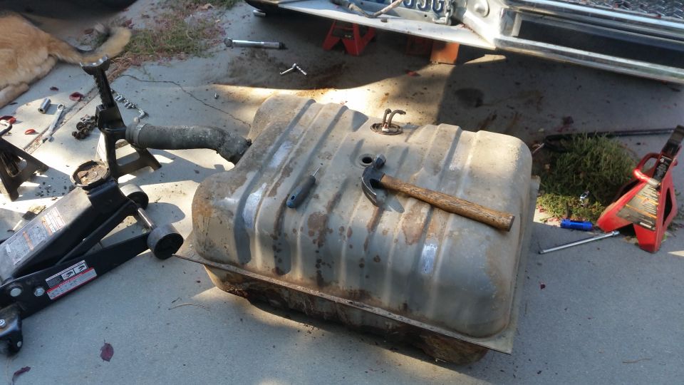 Rusty fuel tank on the ground next to car jack and hammer, with vehicle in background.