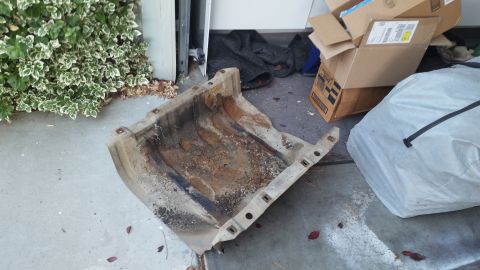 1985 Ford Bronco - A rusty, metal skid plate lies on a concrete driveway, partially covered in dirt and debris. Cardboard boxes and a large white tarp are vis…