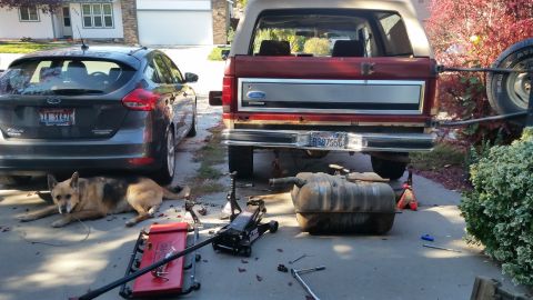 1985 Ford Bronco - The rear of a red Ford Bronco is shown with its fuel tank removed and resting on the ground. A dark grey Ford Focus is parked to the left,…