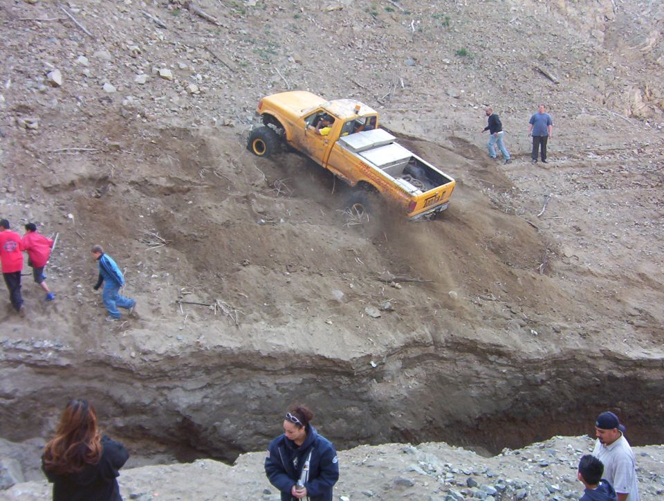 Yellow lifted pickup climbing a steep dirt hill at an off-road park while spectators watch